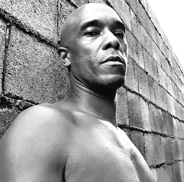 Retrato em preto e branco de Marcus Chocollate encostado num muro de blocos.